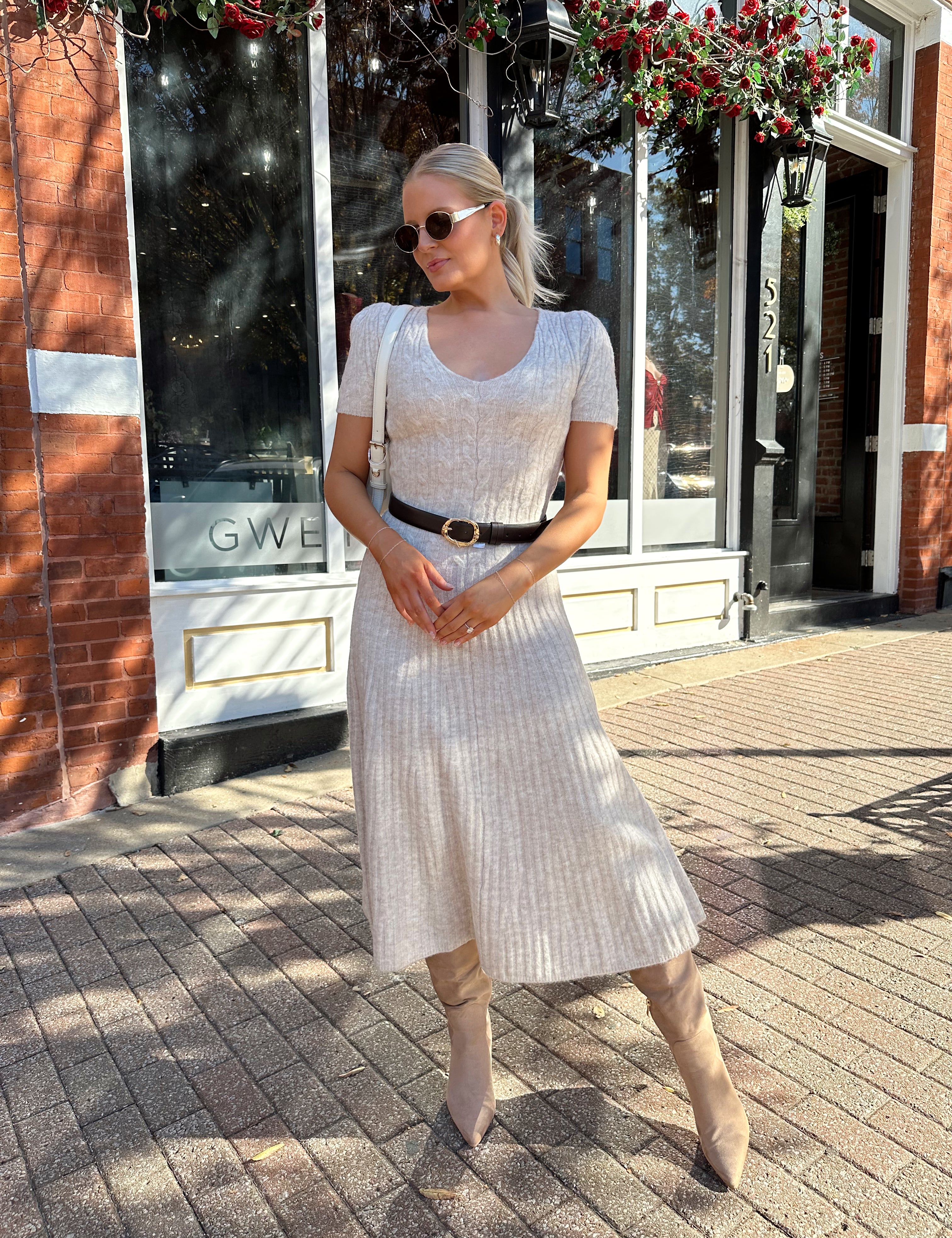 Woman wearing oatmeal cable knit sweater dress with dark belt. 
