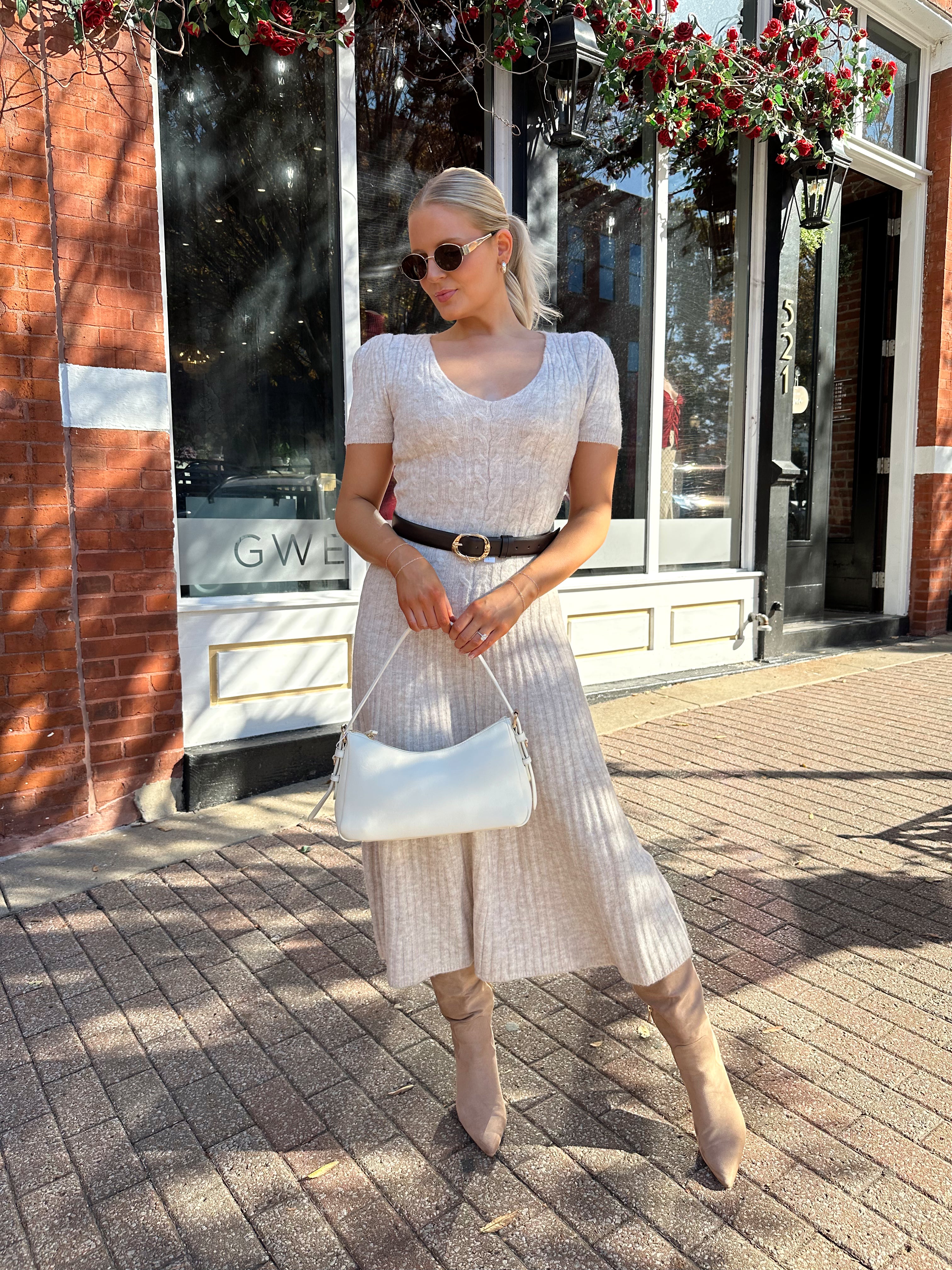 Woman wearing oatmeal cable knit sweater dress with dark belt. 