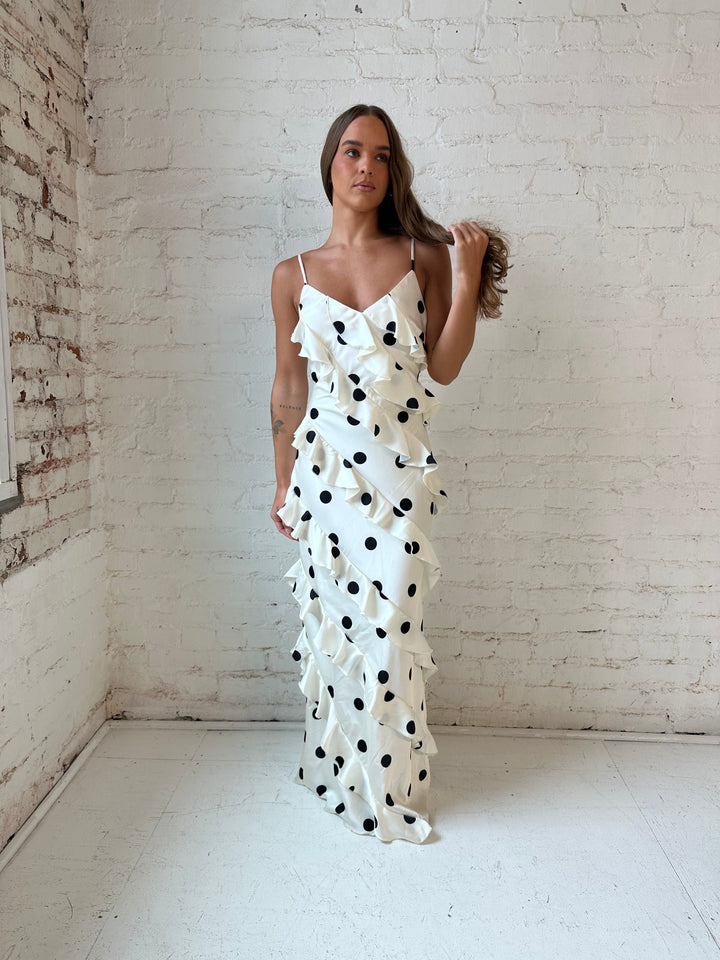 Woman wearing cream and black polka dot ruffle maxi dress with side slit.