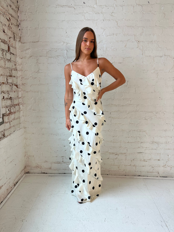 Woman wearing cream and black polka dot ruffle maxi dress with side slit.
