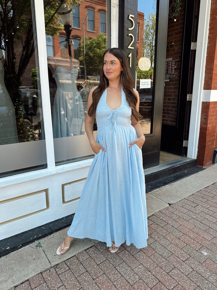 Woman wearing blue smocked midi dress with halter neck, tie detail, and pockets.