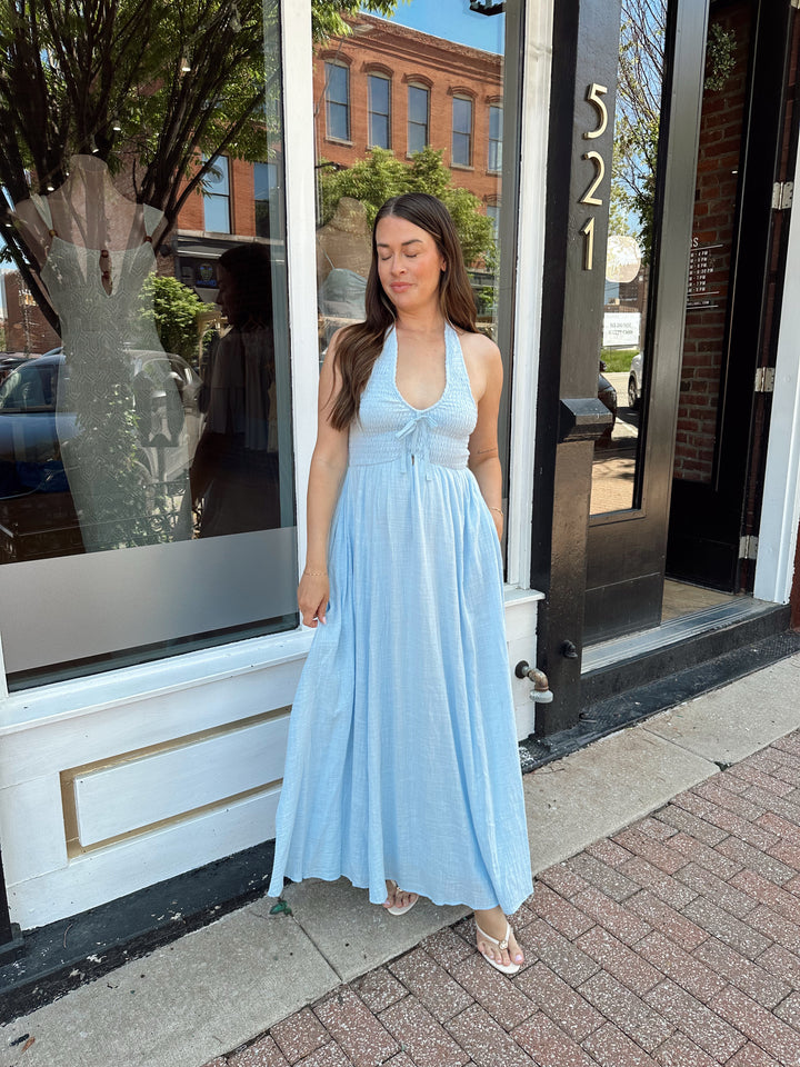 Woman wearing blue smocked midi dress with halter neck, tie detail, and pockets.