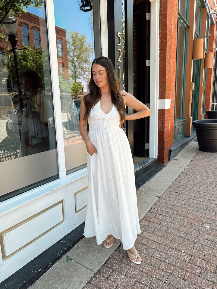 Woman wearing white smocked midi dress with a halter neck, tie detail, and pockets.
