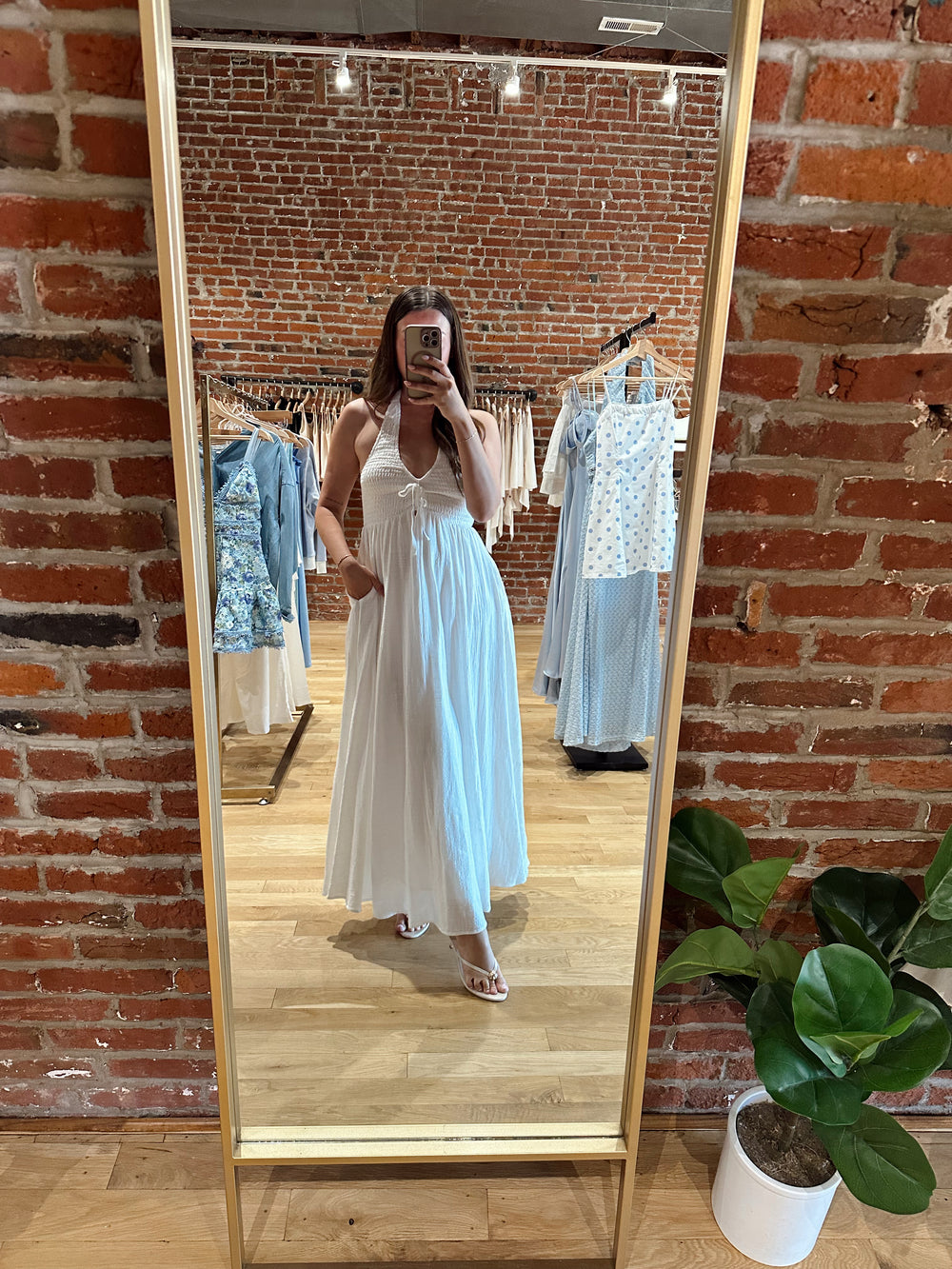 Woman wearing white smocked midi dress with a halter neck, tie detail, and pockets.