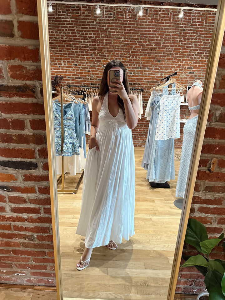 Woman wearing white smocked midi dress with a halter neck, tie detail, and pockets.