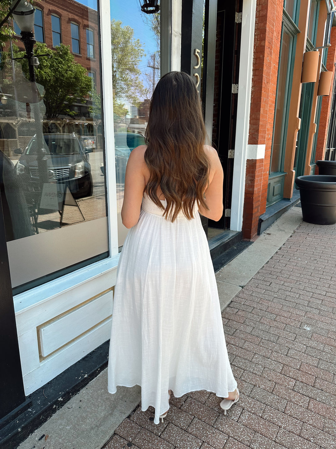 Woman wearing white smocked midi dress with a halter neck, tie detail, and pockets.