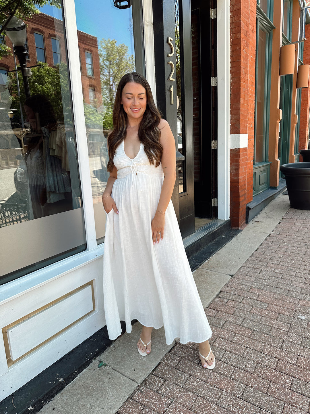 Woman wearing white smocked midi dress with a halter neck, tie detail, and pockets.
