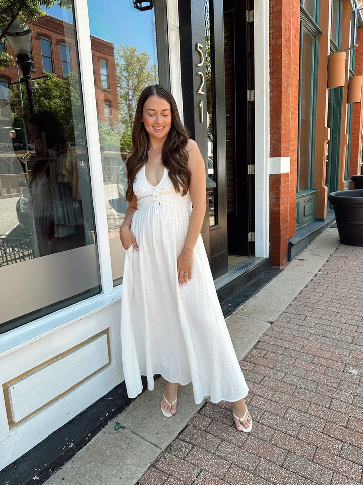 Woman wearing white smocked midi dress with a halter neck, tie detail, and pockets.