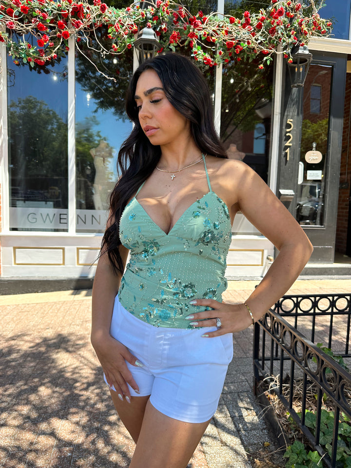 Woman wearing sage green beaded halter neck top. V-neckline, open back, and white micro shorts. 