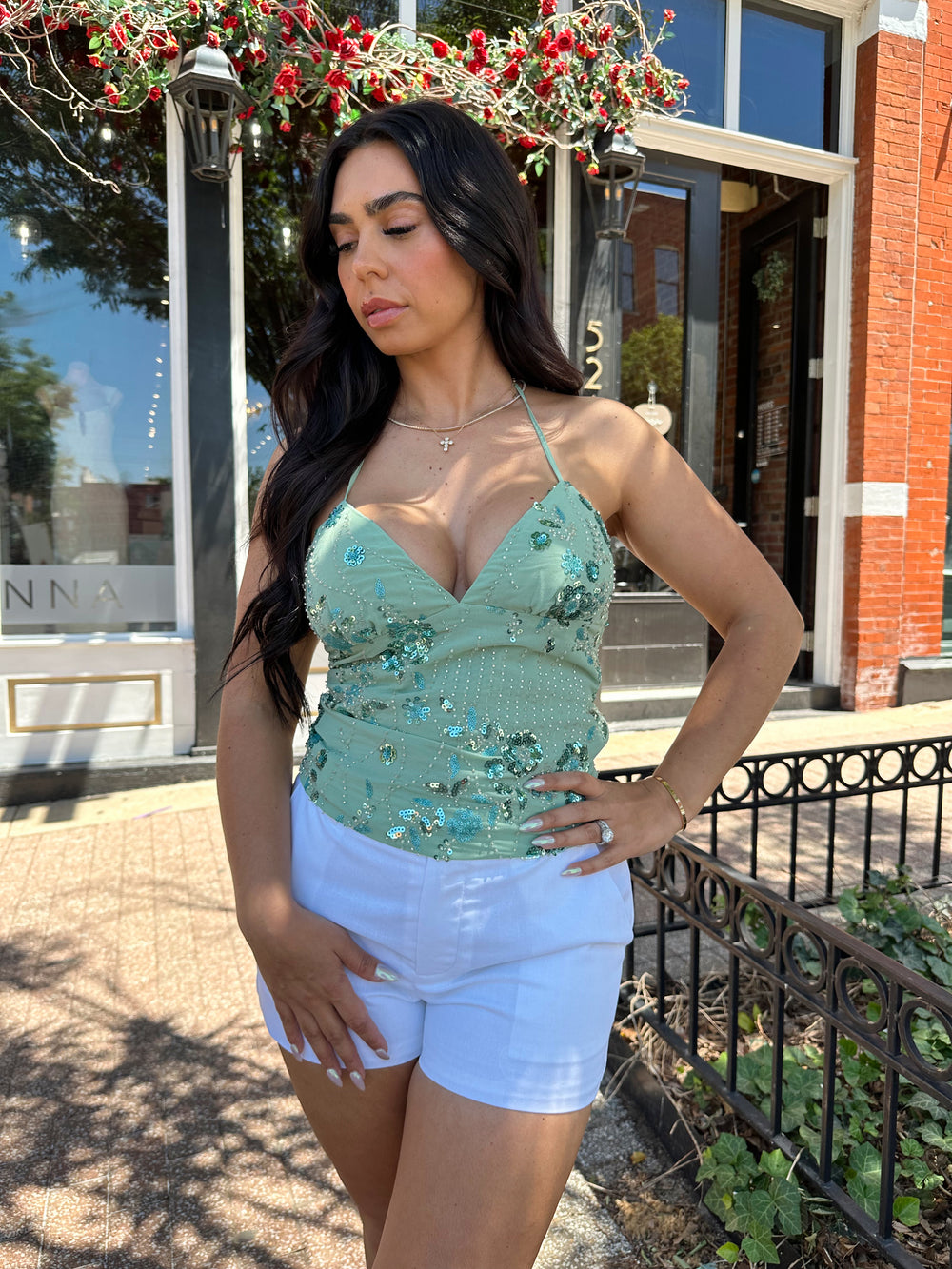 Woman wearing sage green beaded halter neck top. V-neckline, open back, and white micro shorts. 