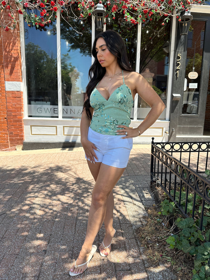 Woman wearing sage green beaded halter neck top. V-neckline, open back, and white micro shorts. 