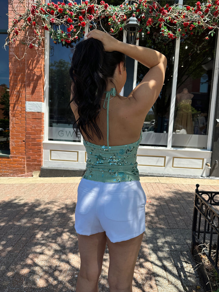 Woman wearing sage green beaded halter neck top. V-neckline, open back, and white micro shorts. 