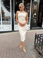 Woman wearing tan halter midi dress with gold chain link detail and slit up the back.