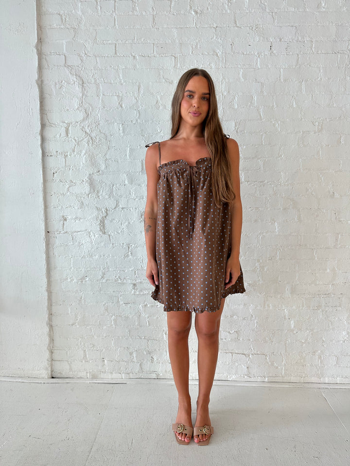 Woman wearing brown babydoll style mini dress with blue polka dots and self tie straps. 