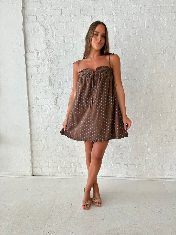Woman wearing brown babydoll style mini dress with blue polka dots and self tie straps. 