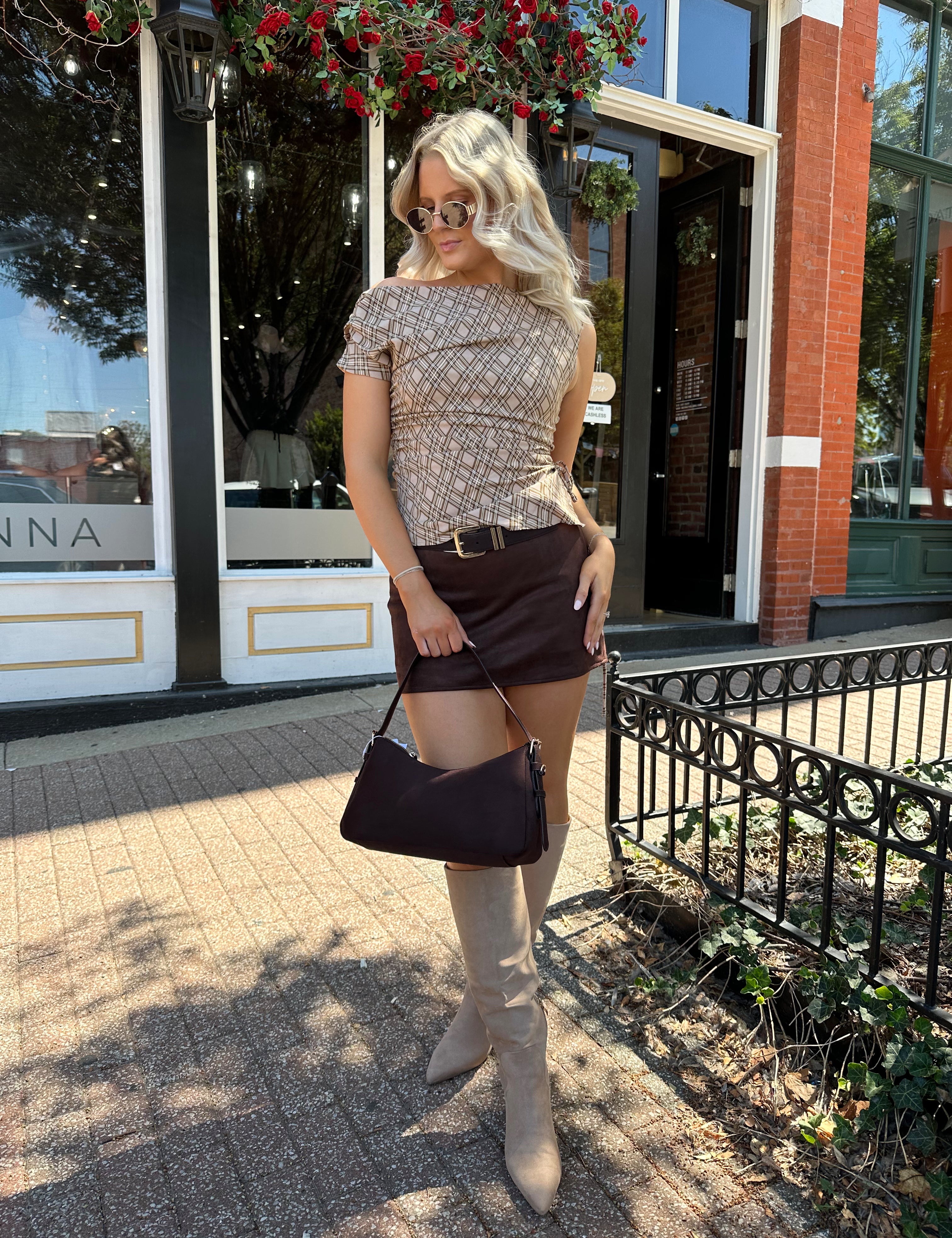 Woman wearing brown plaid asymmetrical top with adjustable side cutout and brown suede mini skirt. 