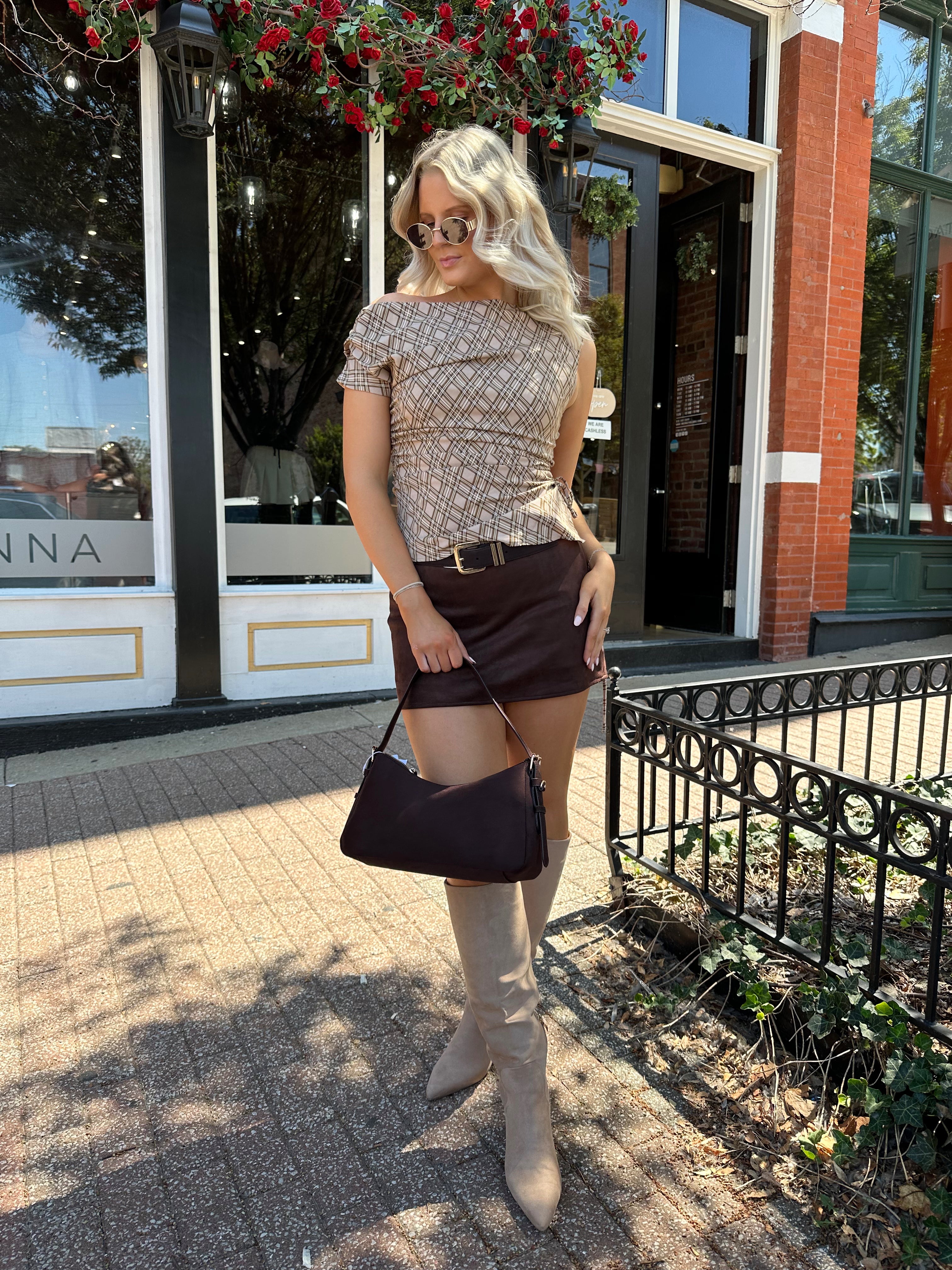Woman wearing brown plaid asymmetrical top with adjustable side cutout and brown suede mini skirt. 