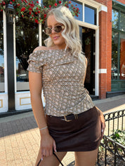 Woman wearing brown plaid asymmetrical top with adjustable side cutout and brown suede mini skirt. 