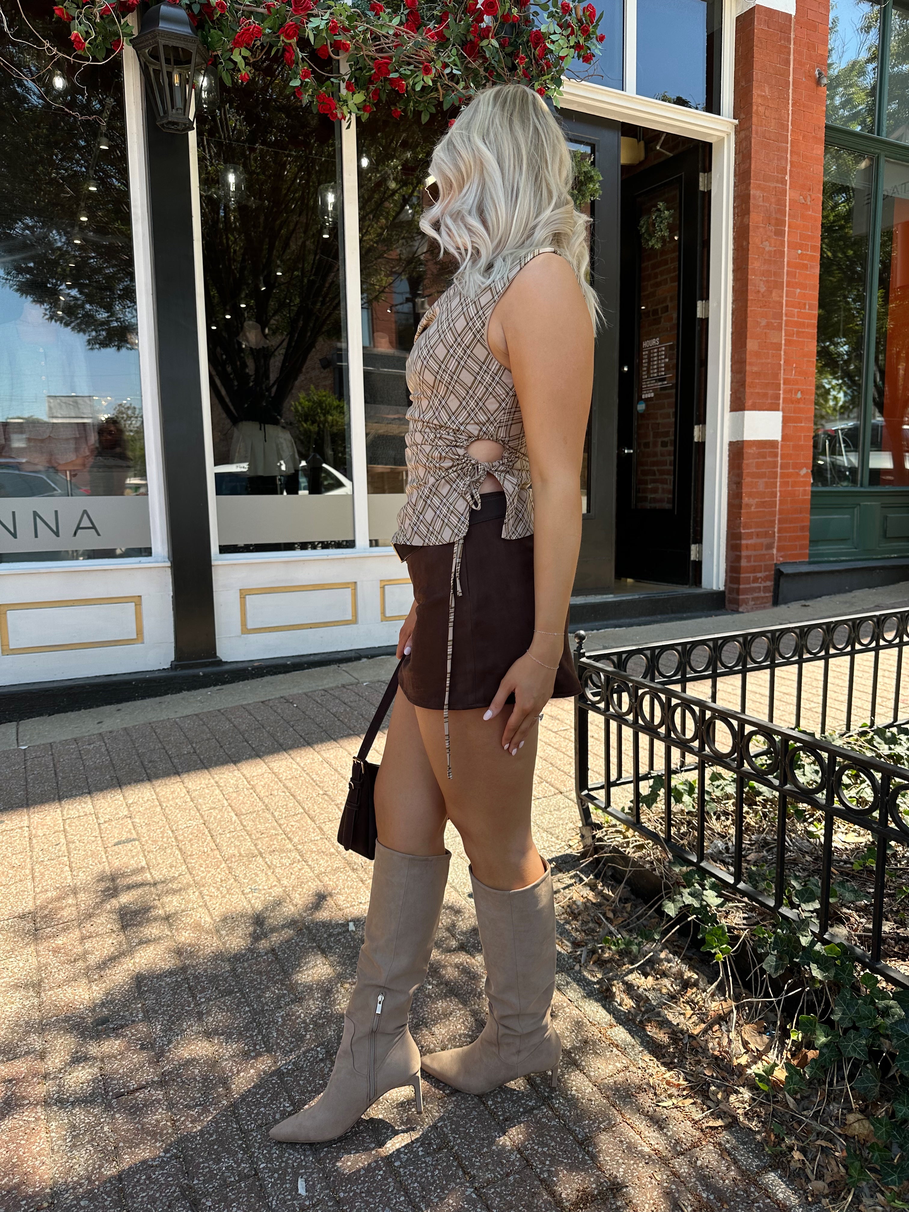 Woman wearing brown plaid asymmetrical top with adjustable side cutout and brown suede mini skirt. 