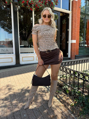 Woman wearing brown plaid asymmetrical top with adjustable side cutout and brown suede mini skirt. 