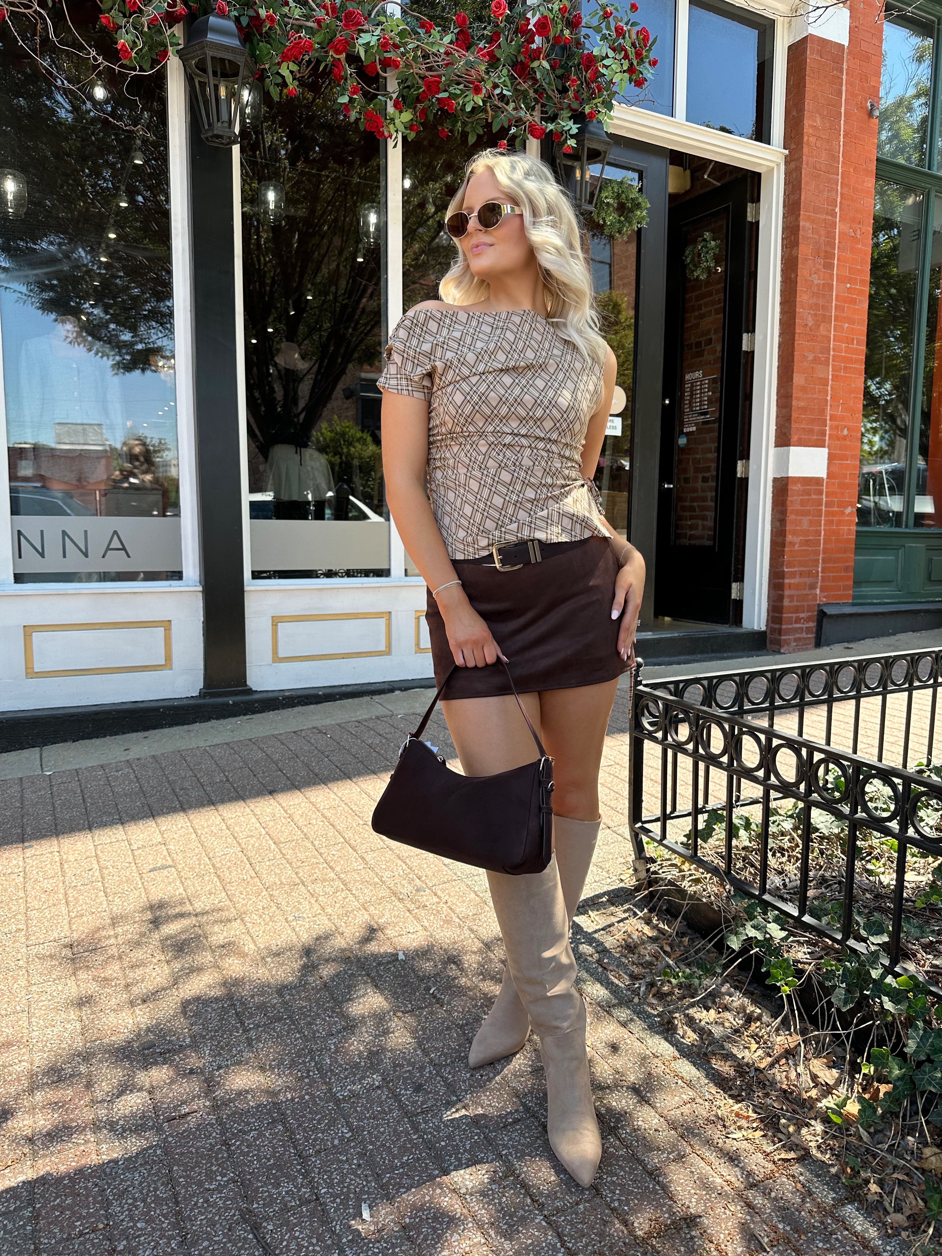 Woman wearing brown plaid asymmetrical top with adjustable side cutout and brown suede mini skirt. 