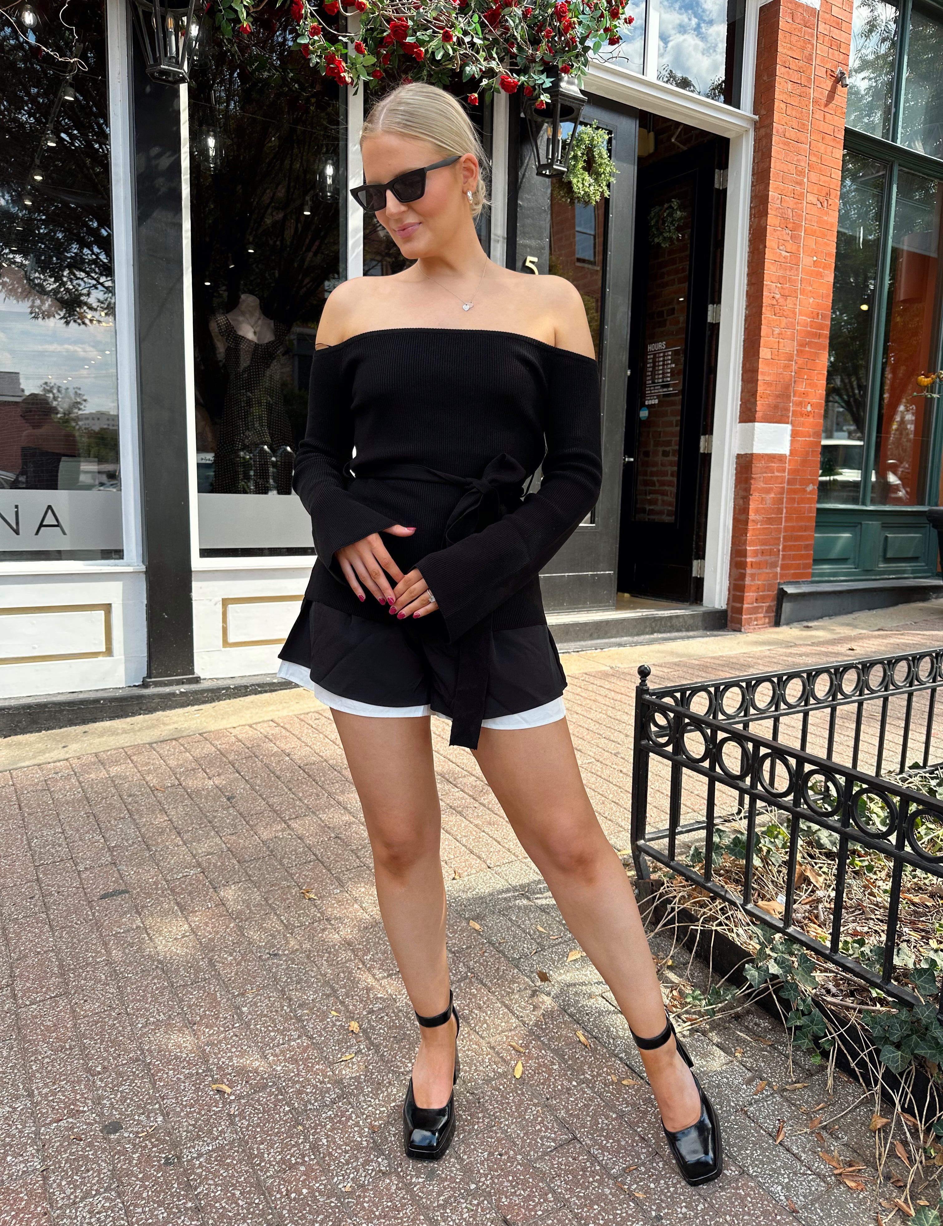 Woman wearing black ribbed off the shoulder top with belt tie detail.