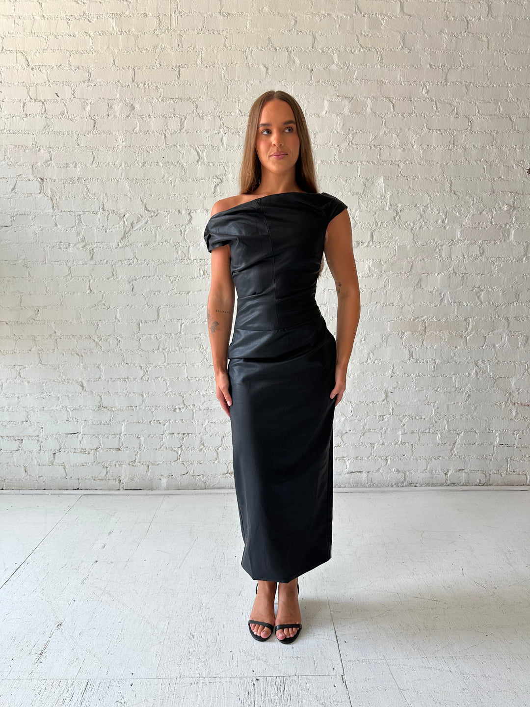 Woman wearing black leather midi dress with an asymmetrical neckline, and back slit.