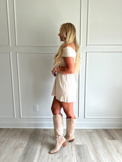 Woman wearing a light beige knit mini dress with an off the shoulder band, and pleated bottom.