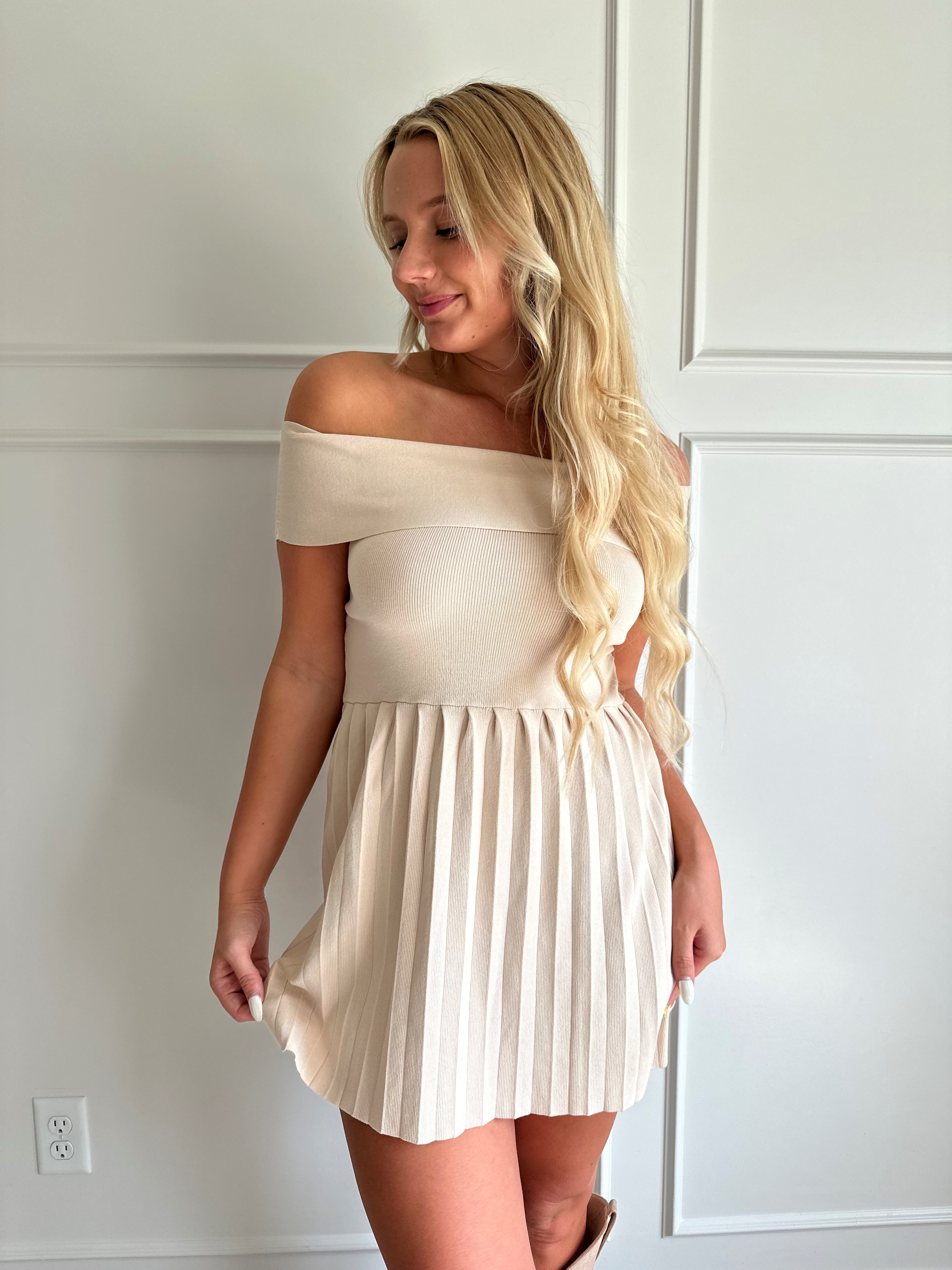 Woman wearing a light beige knit mini dress with an off the shoulder band, and pleated bottom.