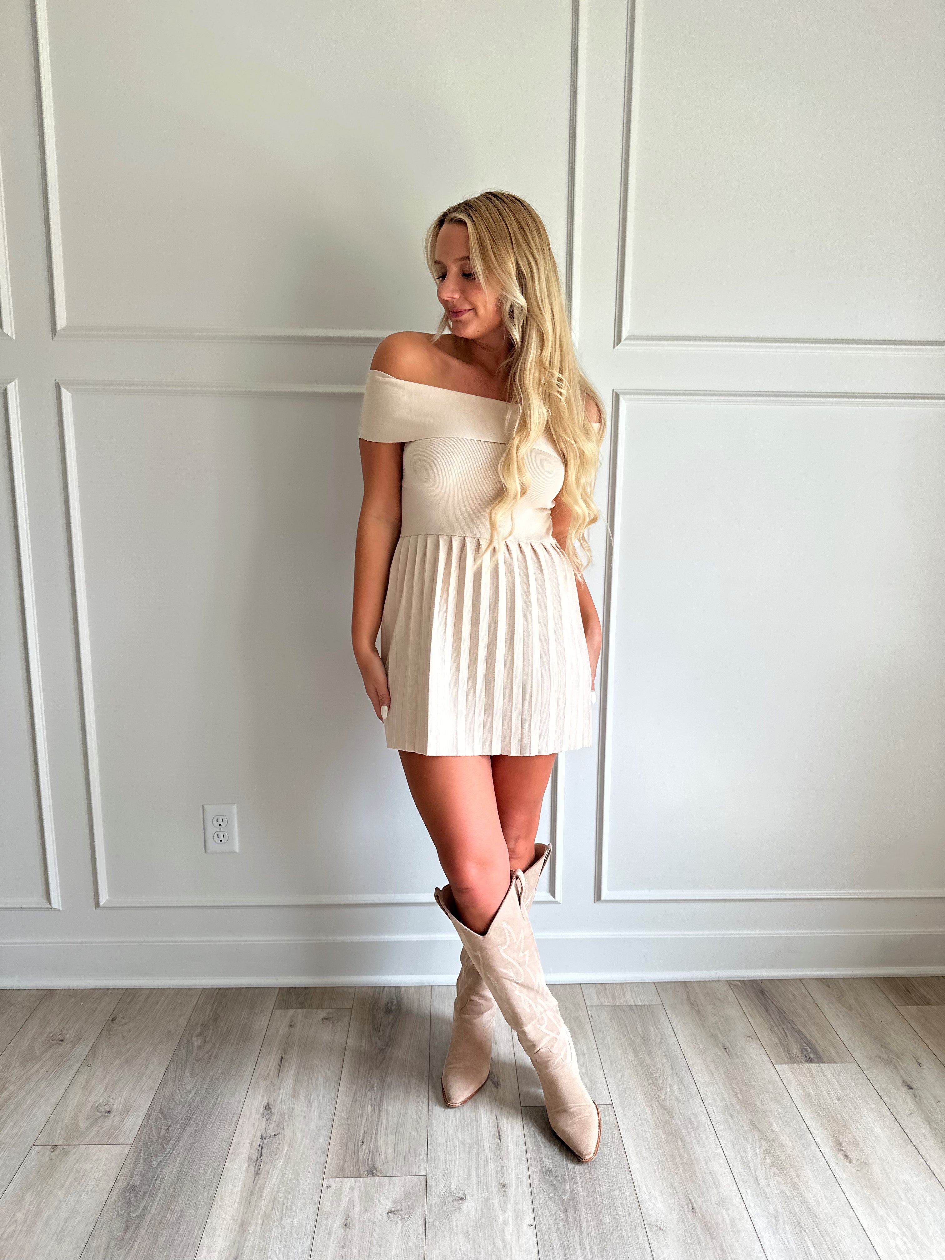 Woman wearing a light beige knit mini dress with an off the shoulder band, and pleated bottom.