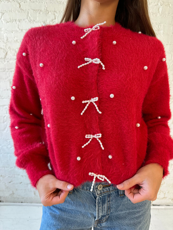 Woman wearing bright red knit cardigan with pearl accents, and pearl bow button closure. 