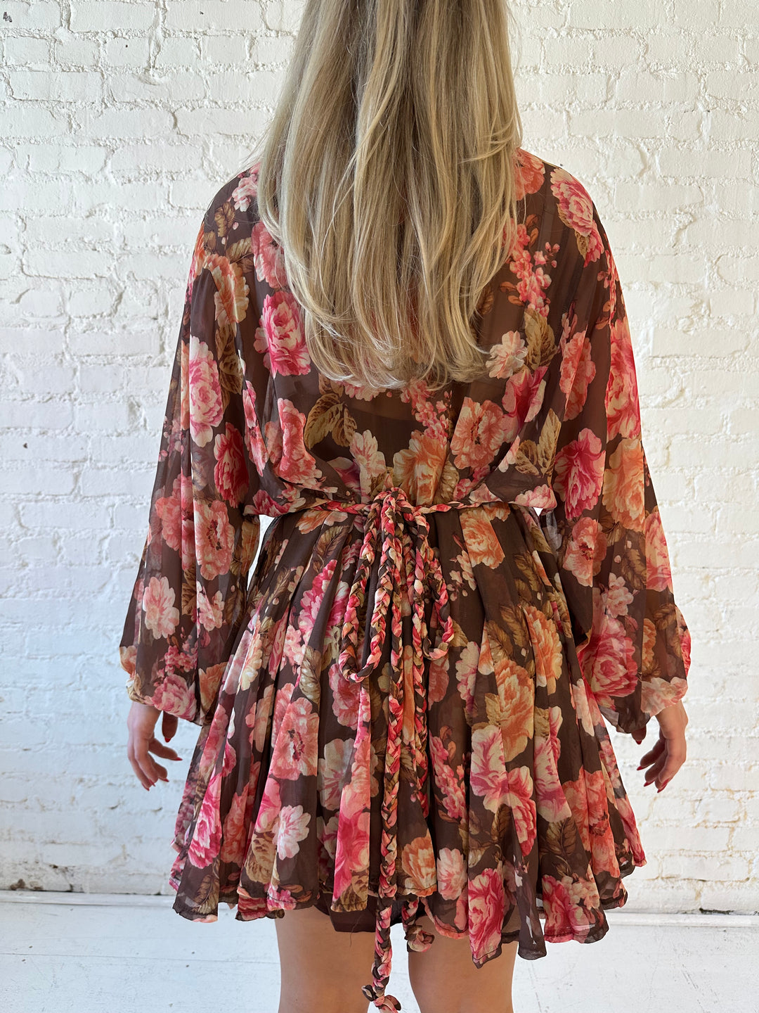 Woman wearing pink and brown floral long sleeve flouncey mini dress with braided waist tie. 
