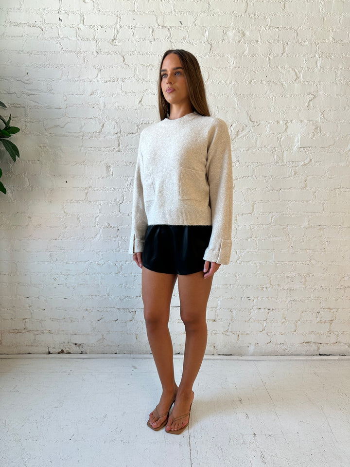 Woman wearing taupe knit sweater with front pockets, little wrist detailing, and faux leather shorts. 