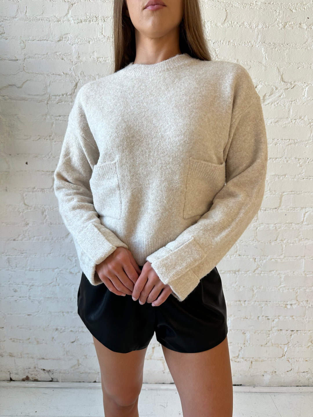 Woman wearing taupe knit sweater with front pockets, little wrist detailing, and faux leather shorts. 