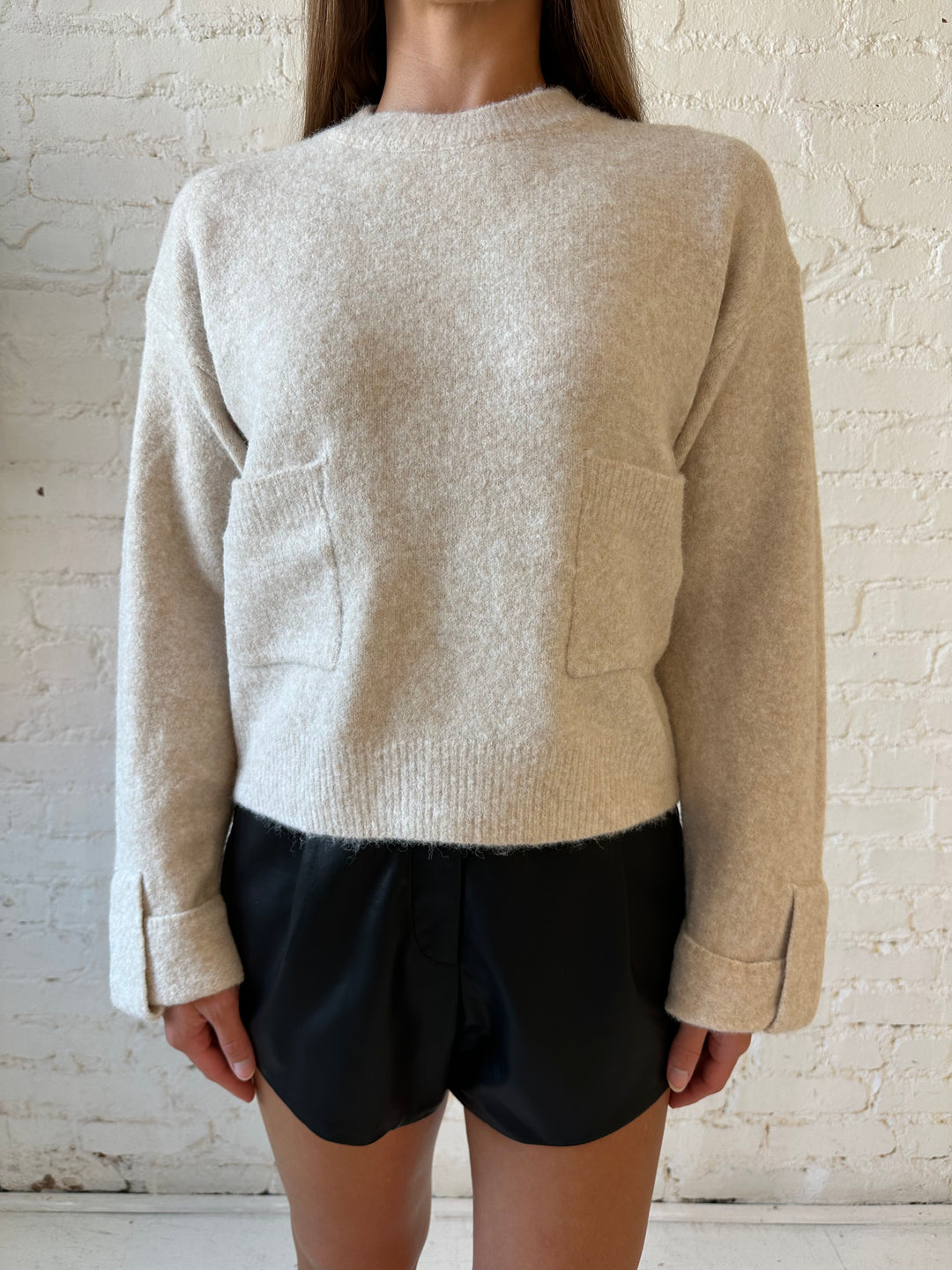 Woman wearing taupe knit sweater with front pockets, little wrist detailing, and faux leather shorts. 