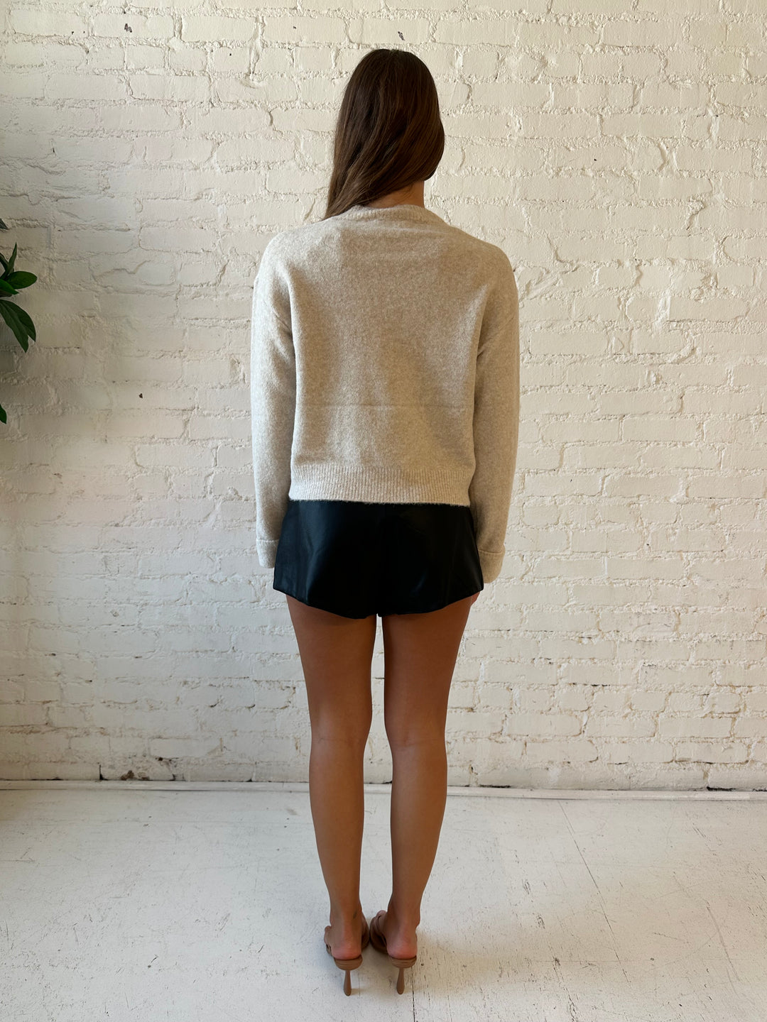 Woman wearing taupe knit sweater with front pockets, little wrist detailing, and faux leather shorts. 