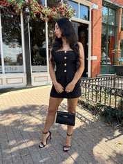Woman wearing black strapless two piece set with mini skort and gold button detailing.