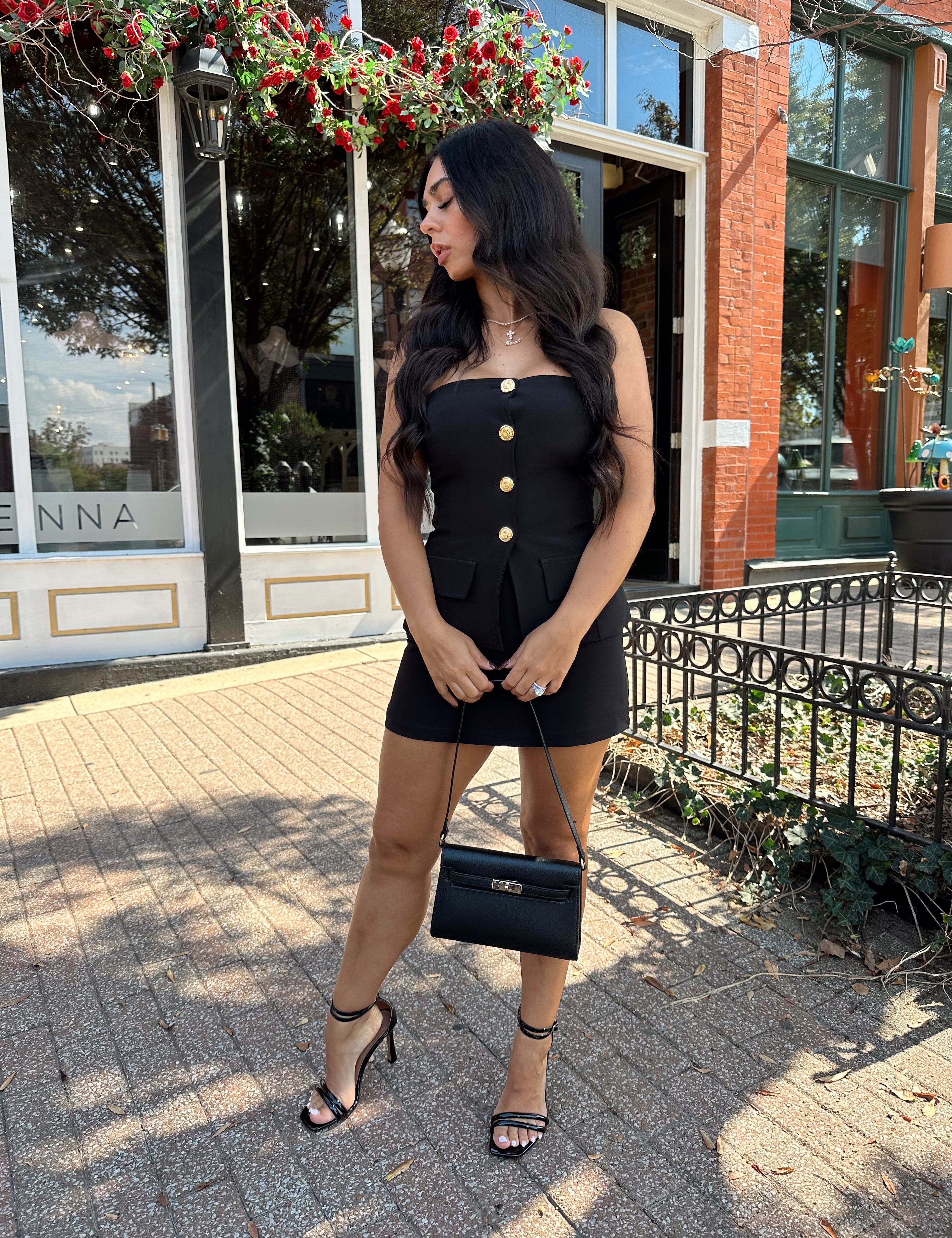 Woman wearing black strapless two piece set with mini skort and gold button detailing.