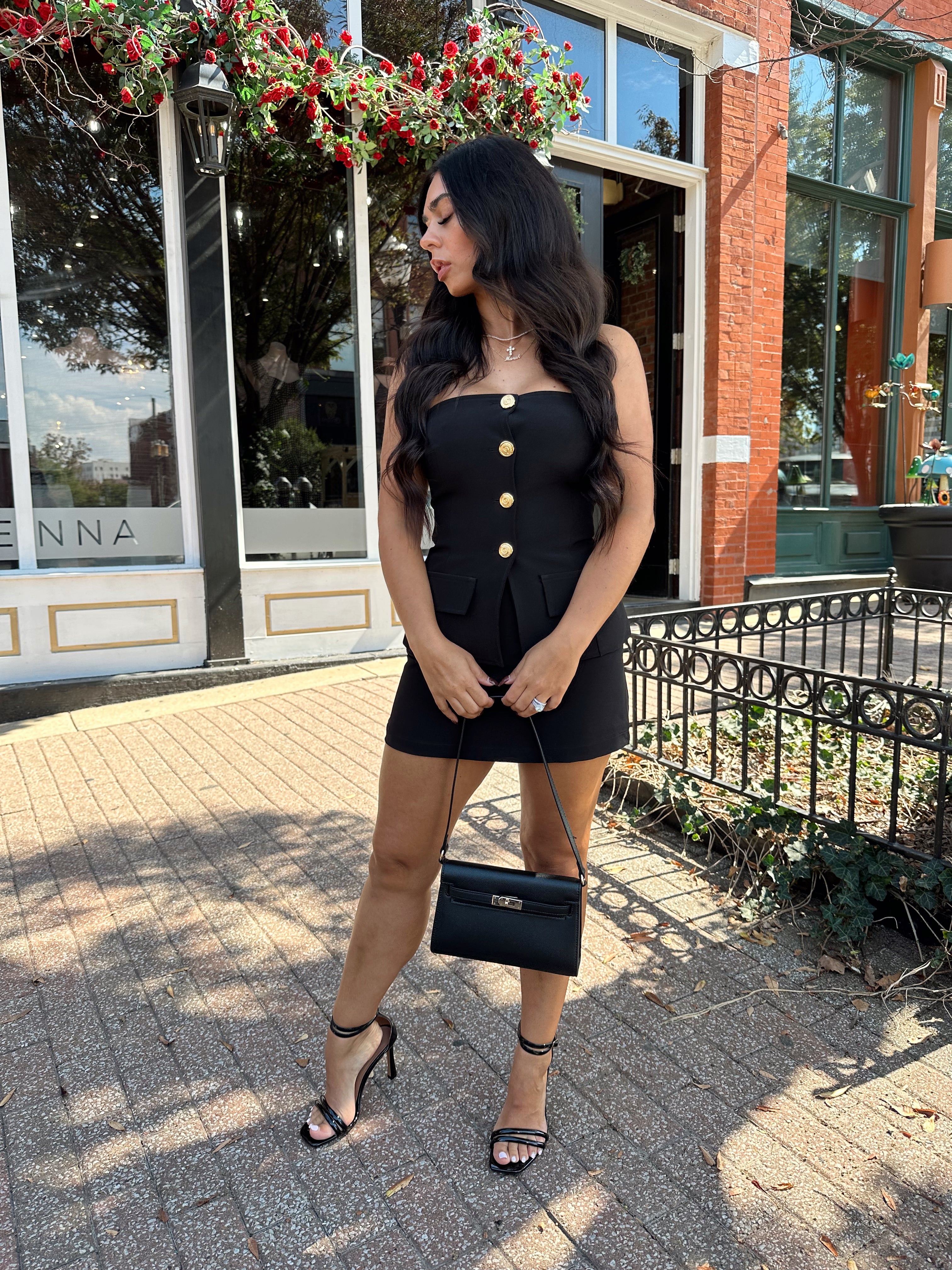 Woman wearing black strapless two piece set with mini skort and gold button detailing.