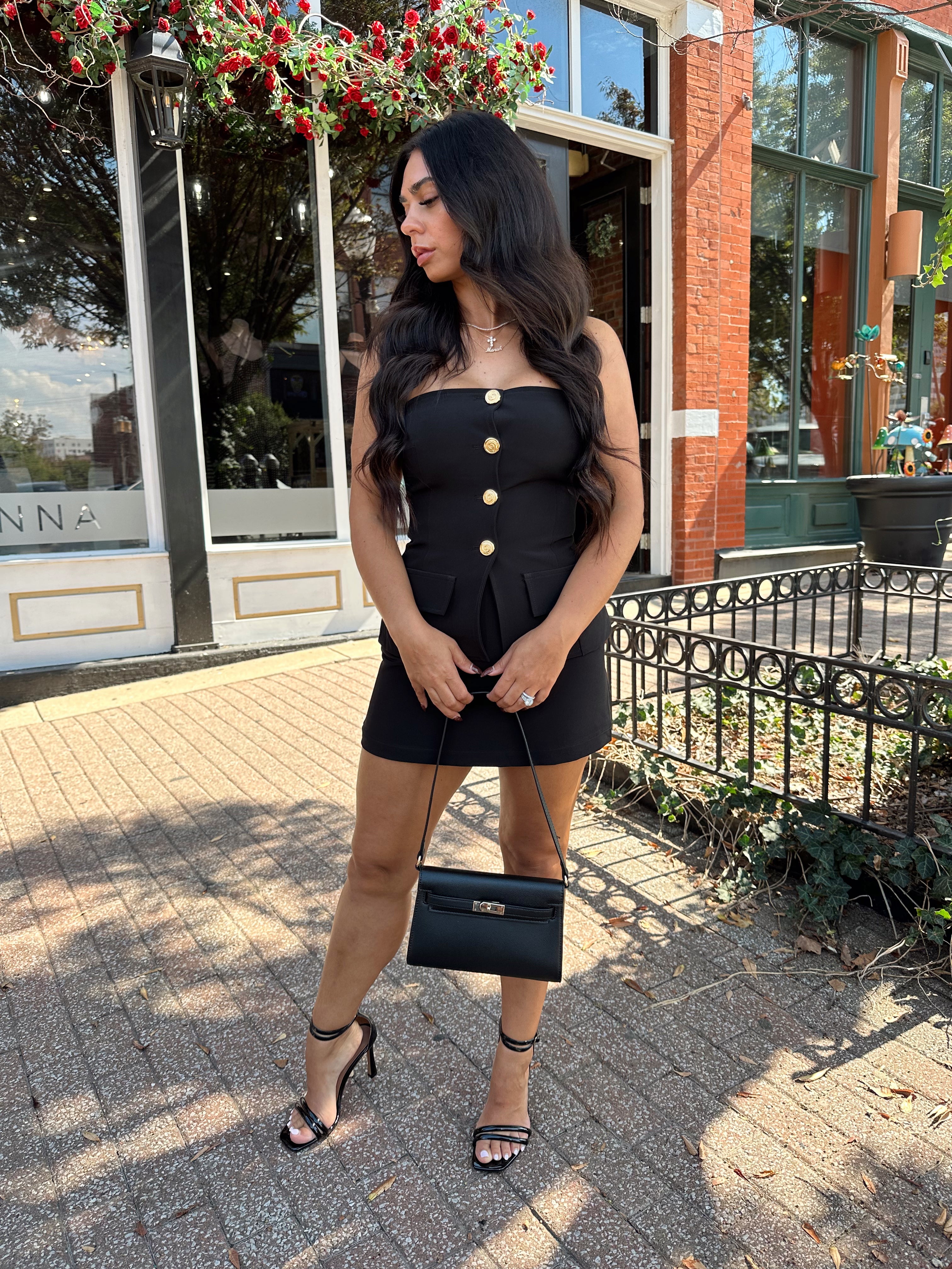 Woman wearing black strapless two piece set with mini skort and gold button detailing.