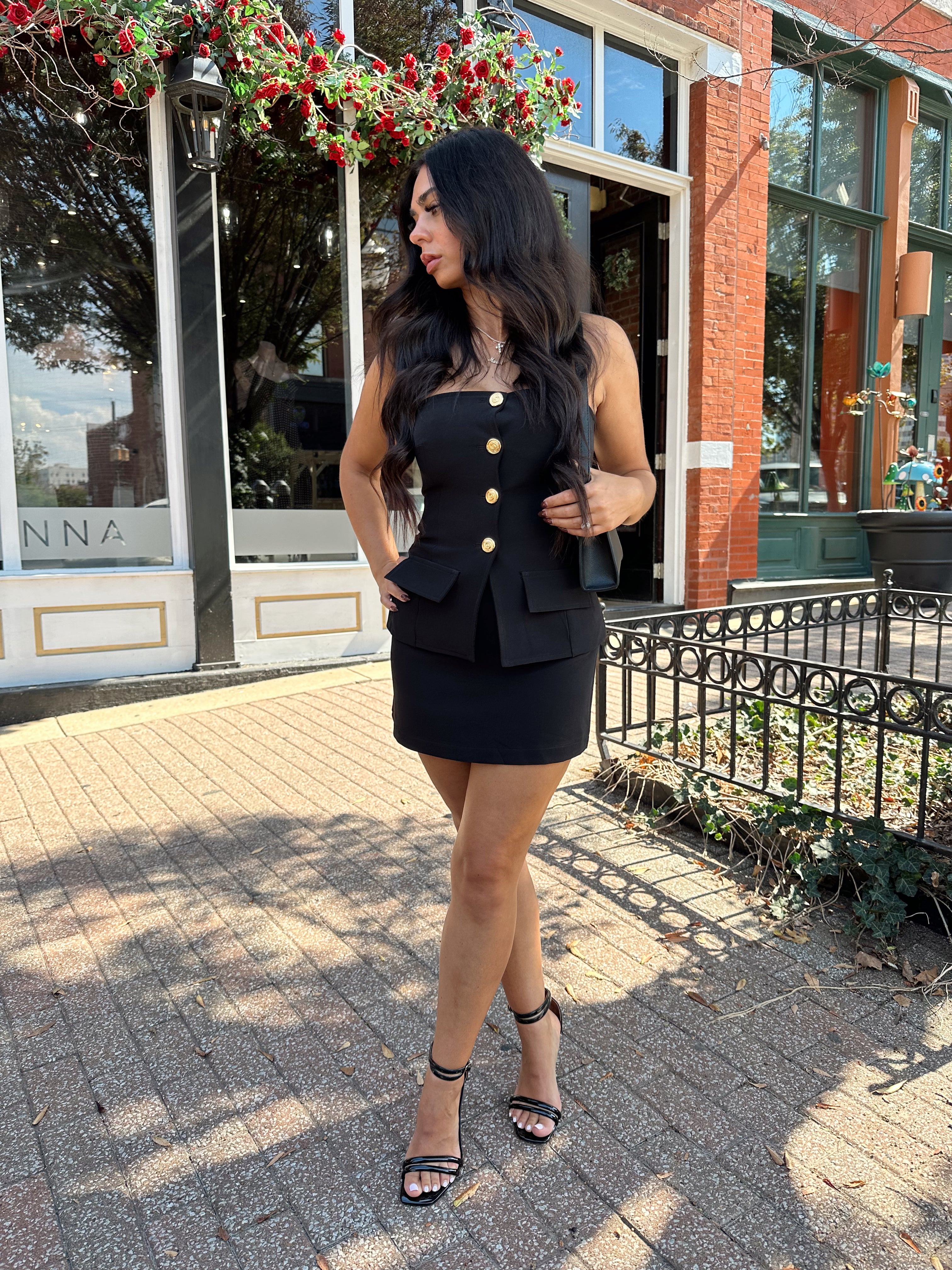 Woman wearing black strapless two piece set with mini skort and gold button detailing.