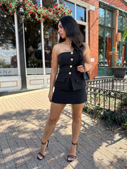Woman wearing black strapless two piece set with mini skort and gold button detailing.