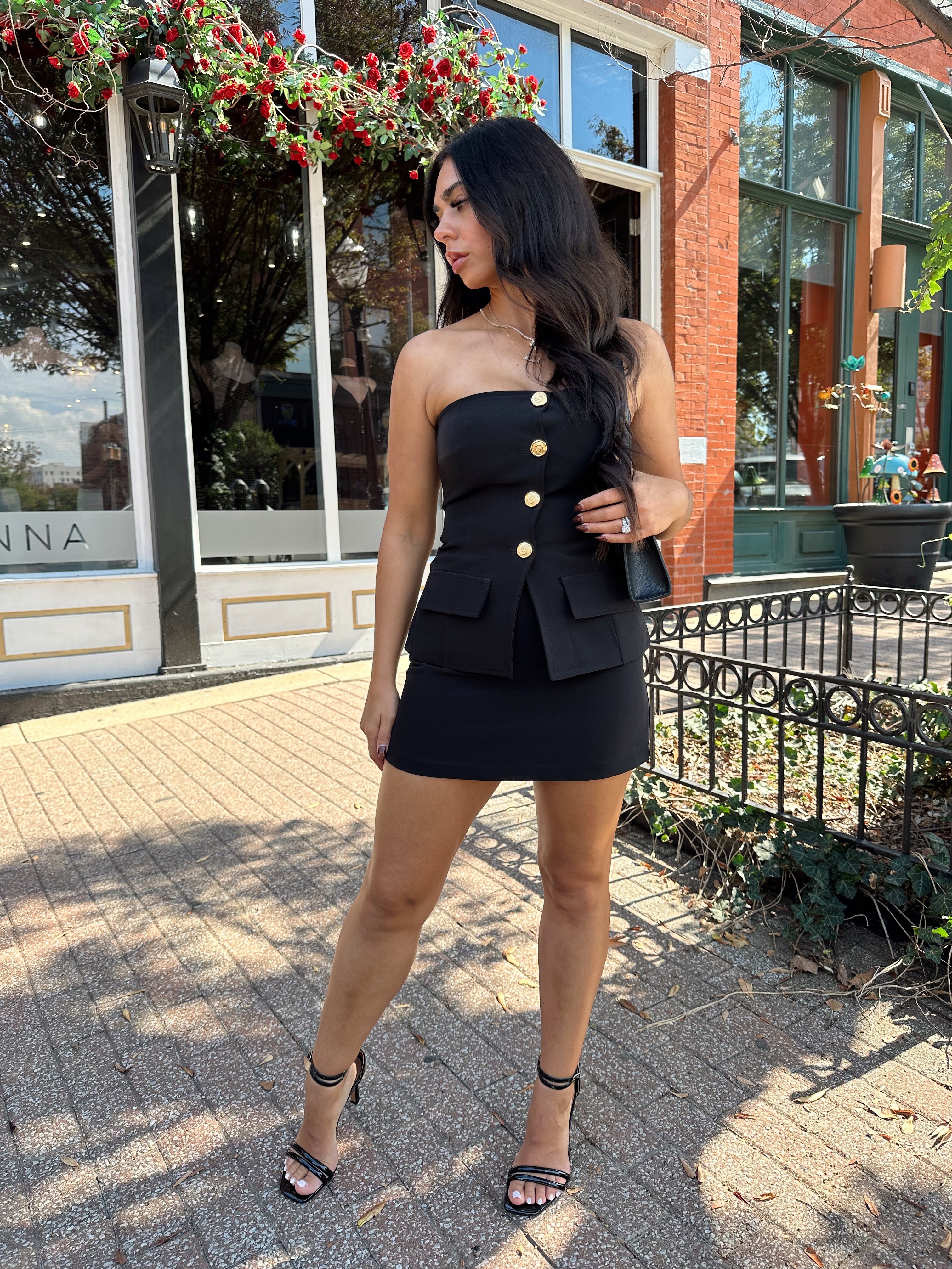 Woman wearing black strapless two piece set with mini skort and gold button detailing.