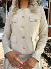 Woman wearing a light beige faux leather jacket with gold button closure. 