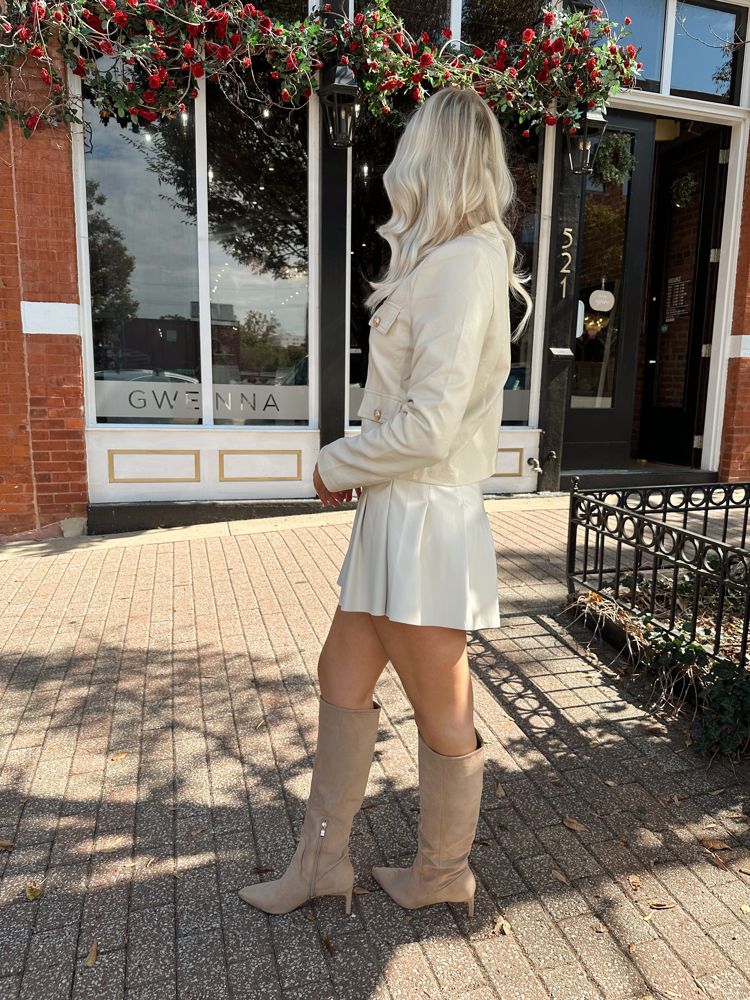 Woman wearing a light beige faux leather jacket with gold button closure. 