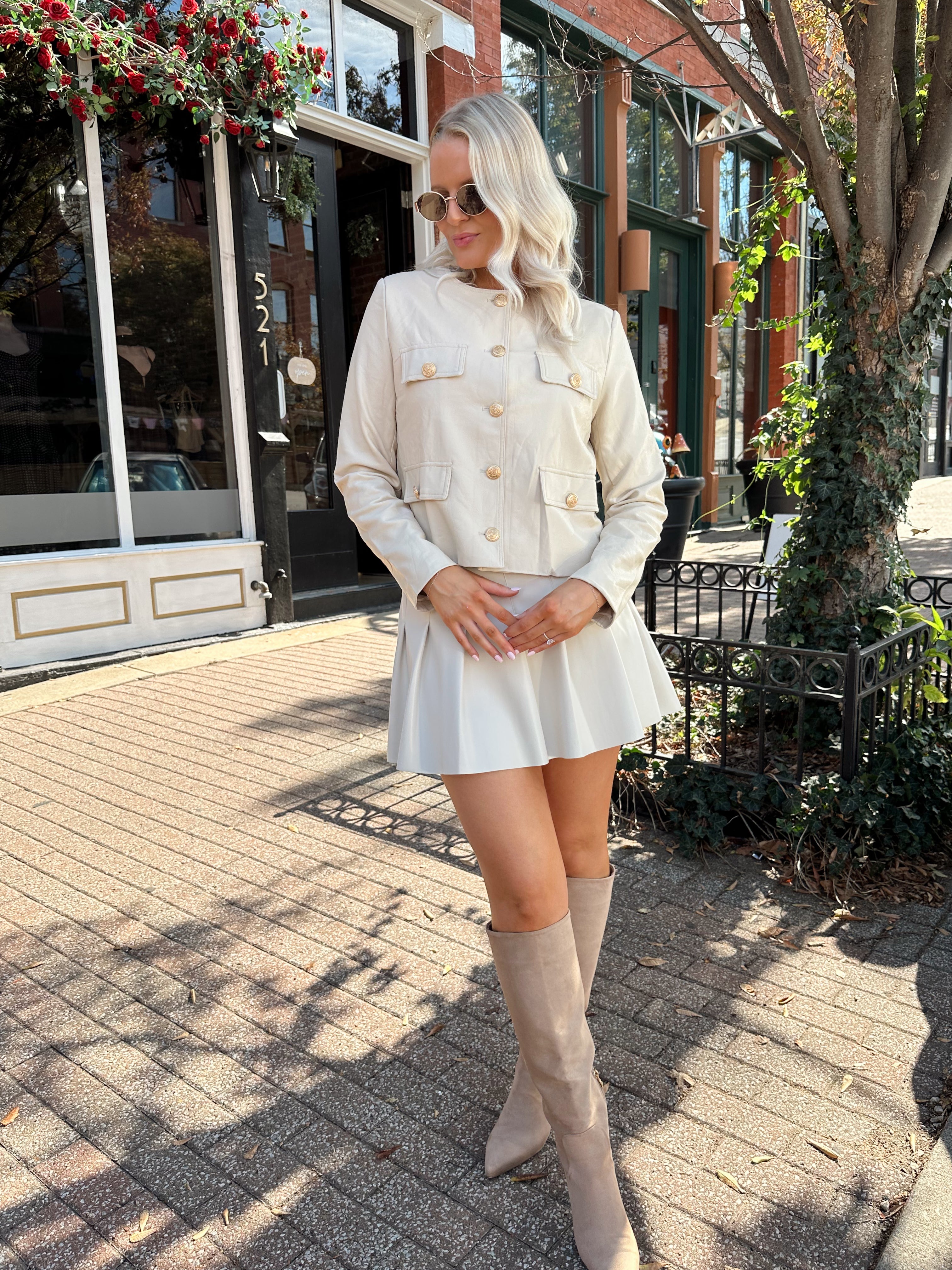 Woman wearing a light beige faux leather jacket with gold button closure. 