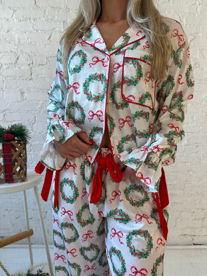 Woman wearing white satin pajama set with green wreaths all over, and red velvet trim. 