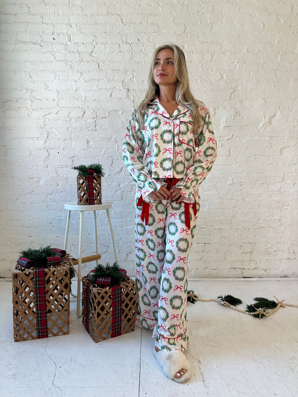 Woman wearing white satin pajama set with green wreaths all over, and red velvet trim. 
