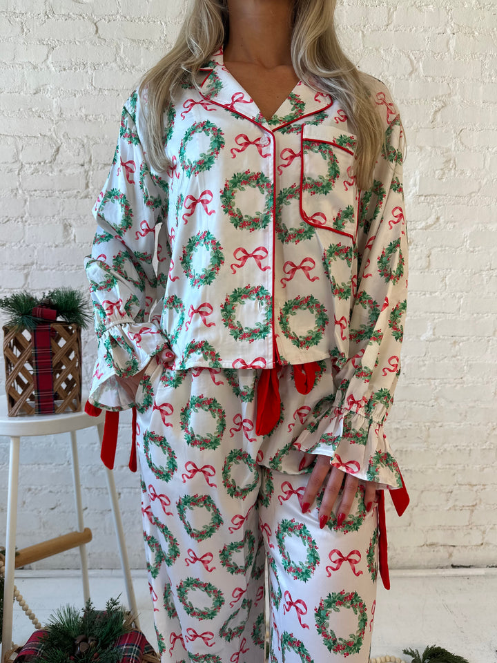 Woman wearing white satin pajama set with green wreaths all over, and red velvet trim. 