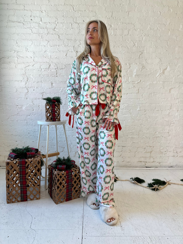 Woman wearing white satin pajama set with green wreaths all over, and red velvet trim. 